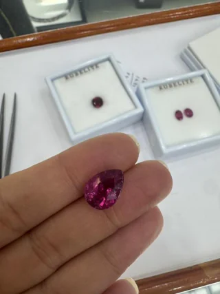 A pear cut rubellite held in the fingers beside labelled stone boxes